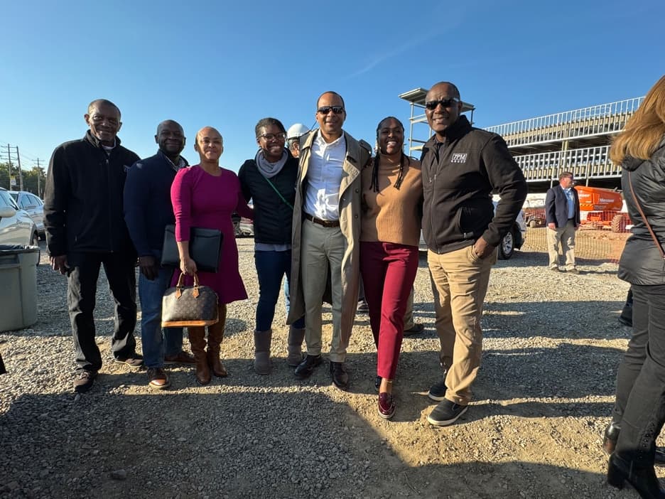 Seven people in business and casual dress stand together on a gravel construction site under a clear sky, with a parking structure under construction behind them