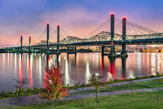 Cable-stayed and truss bridges spanning the Ohio River at dusk, their red-lit towers reflected in the water, with the Louisville skyline behind
