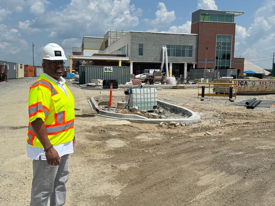 A man in a white hard hat and high-visibility vest stands on the graded dirt of an active construction site, with the partially built hospital behind him