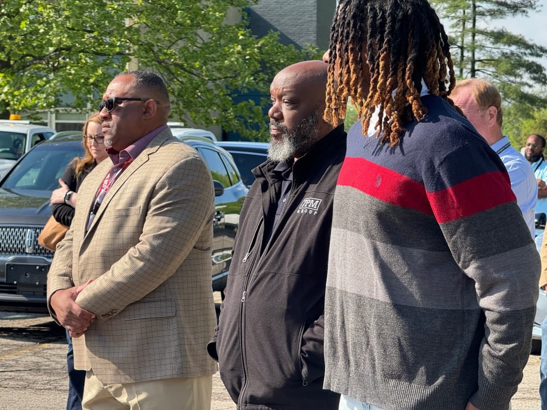 A group of contractors and project staff, including a man in a 7PM Group jacket, standing together in a sunlit parking lot at an outdoor site gathering